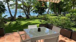 Une terrasse avec une table en verre et des chaises blanches, donnant sur un espace verdoyant avec des arbres et offrant une vue sur la mer.