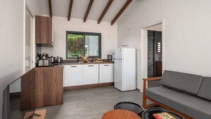A compact kitchen with white cabinets, a refrigerator, and a window with greenery outside. There's a gray sofa and a small round table with a book.