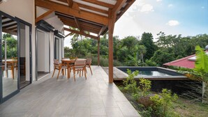 A covered patio with a dining table and chairs, a swimming pool, and a view of lush greenery.