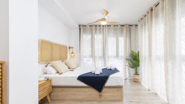 Une chambre à coucher avec un lit, des tables de chevet, un ventilateur de plafond, une plante en pot et des rideaux légers.