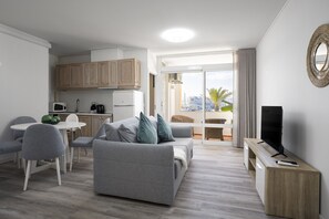 A modern living room with a grey sofa, a dining table with chairs, a kitchen area, and a balcony with a view.