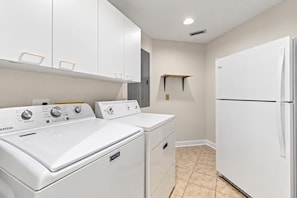 A laundry room with two Maytag washers, a white refrigerator, and a small shelf.