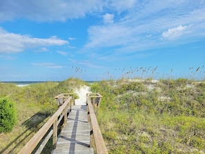 A wooden boardwalk leading to a beach with sand dunes and grass.