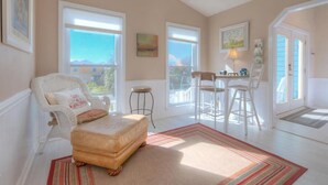 Ein gemütliches Wohnzimmer mit einem weißen Sessel, einem kleinen Hocker, einem Esstisch mit Stühlen und Blick ins Freie durch große Fenster.