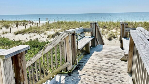 A wooden boardwalk leading to a beach with sand dunes and a clear view of the ocean. - Experience oceanfront luxury at A Waters Edge Escape in Oak Island, NC. (Oak Island)