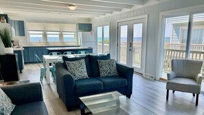 A spacious living area with a sofa, armchair, and coffee table. A kitchen with a dining table and chairs is visible in the background. - Experience oceanfront luxury at A Waters Edge Escape in Oak Island, NC. (Oak Island)