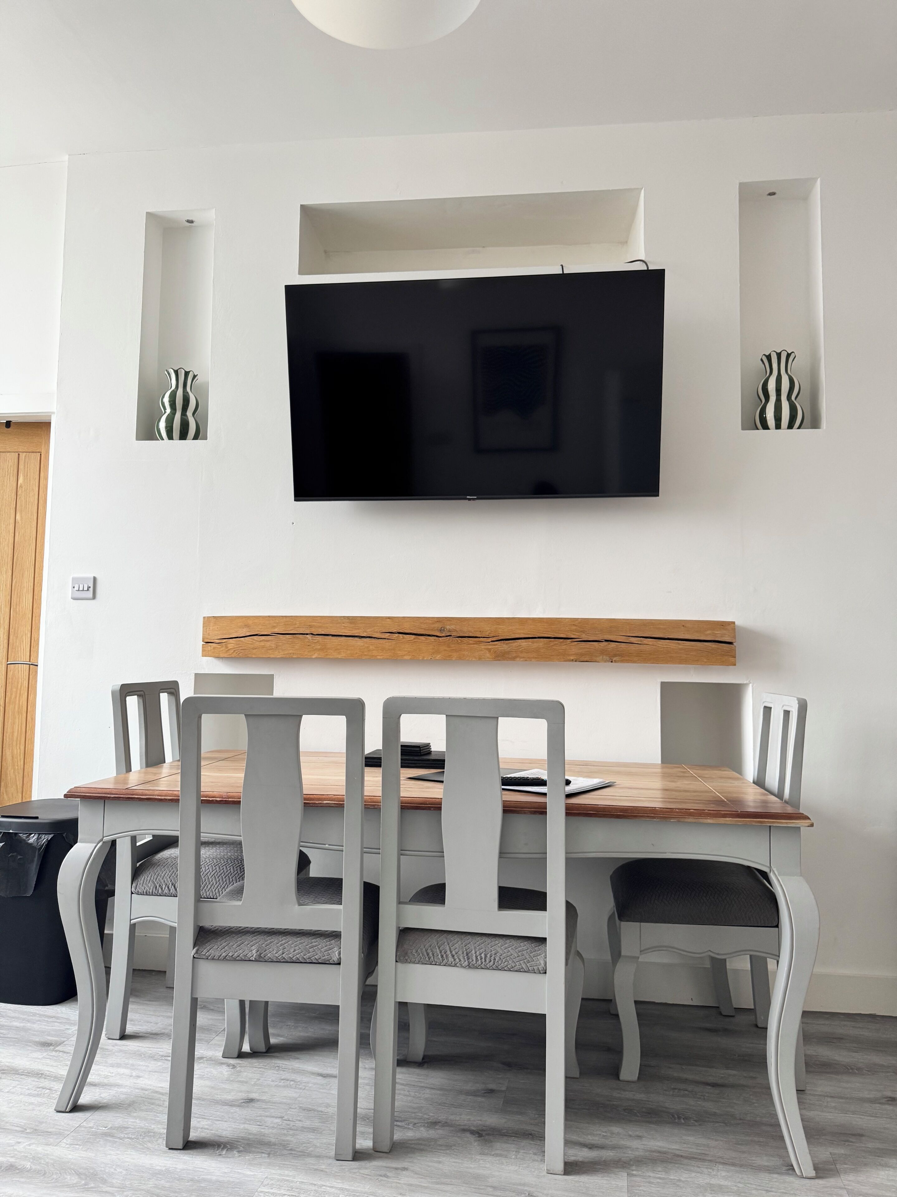 A dining area with a wooden table, grey chairs, a mounted television, and decorative vases.