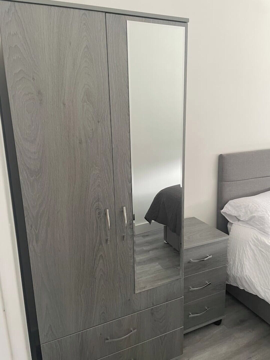 Quarto com guarda-roupa cinzento, cama com roupa de cama branca e cabeceira cinzenta.