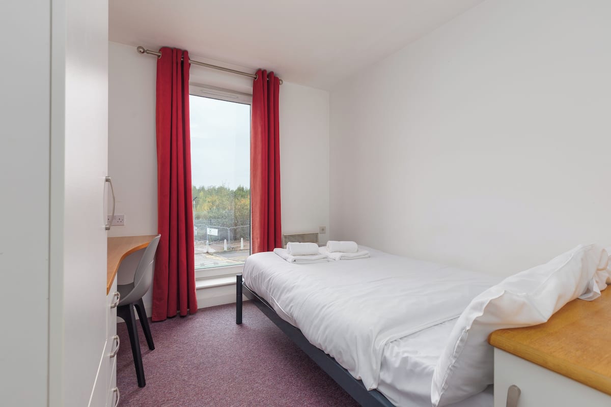 Una camera da letto con un letto, una scrivania e una sedia. Si vede una finestra con tende rosse.