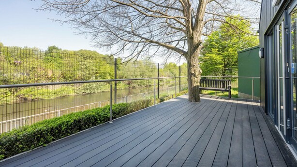 A wooden deck with a metal railing overlooking a river, a tree, and a building with large windows. - Riverside House by UTDM (Salford)