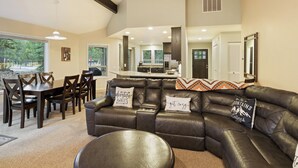 A modern living room with a dark leather sofa, a dining table with chairs, and a kitchen area in the background. - Poplar 32 - Rec Passes for SHARC, Updated home, Centrally located, Private Hot Tub (Sunriver)
