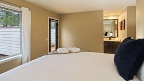 A hotel room with a large bed, a window with curtains, a bathroom with a sink and mirror, and a view of trees outside. - Poplar 32 - Rec Passes for SHARC, Updated home, Centrally located, Private Hot Tub (Sunriver)