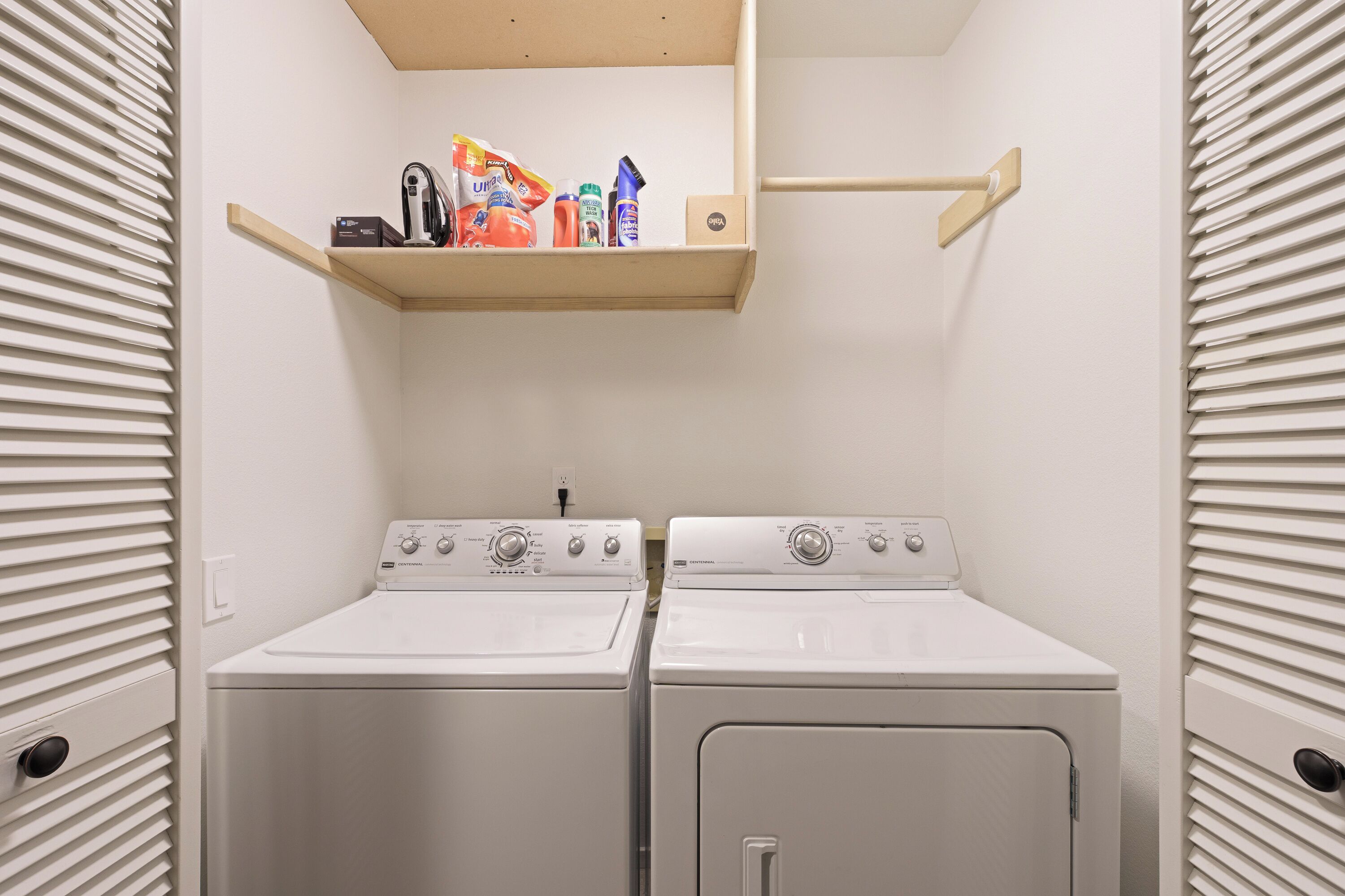 A laundry room with two white front-loading washers and dryers, a wooden shelf above with cleaning supplies, and a wall-mounted towel rack.