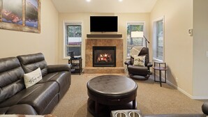 A living room with a fireplace, a leather sofa, a round ottoman, and a flat-screen TV mounted above the fireplace. - Poplar 32 - Rec Passes for SHARC, Updated home, Centrally located, Private Hot Tub (Sunriver)