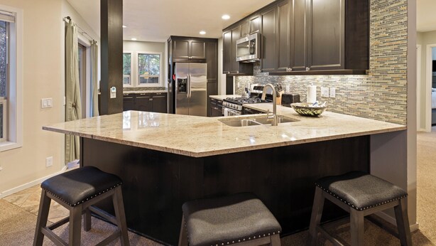 A modern kitchen with dark cabinetry, a large island with stools, stainless steel appliances, and a tiled backsplash. - Poplar 32 - Rec Passes for SHARC, Updated home, Centrally located, Private Hot Tub (Sunriver)