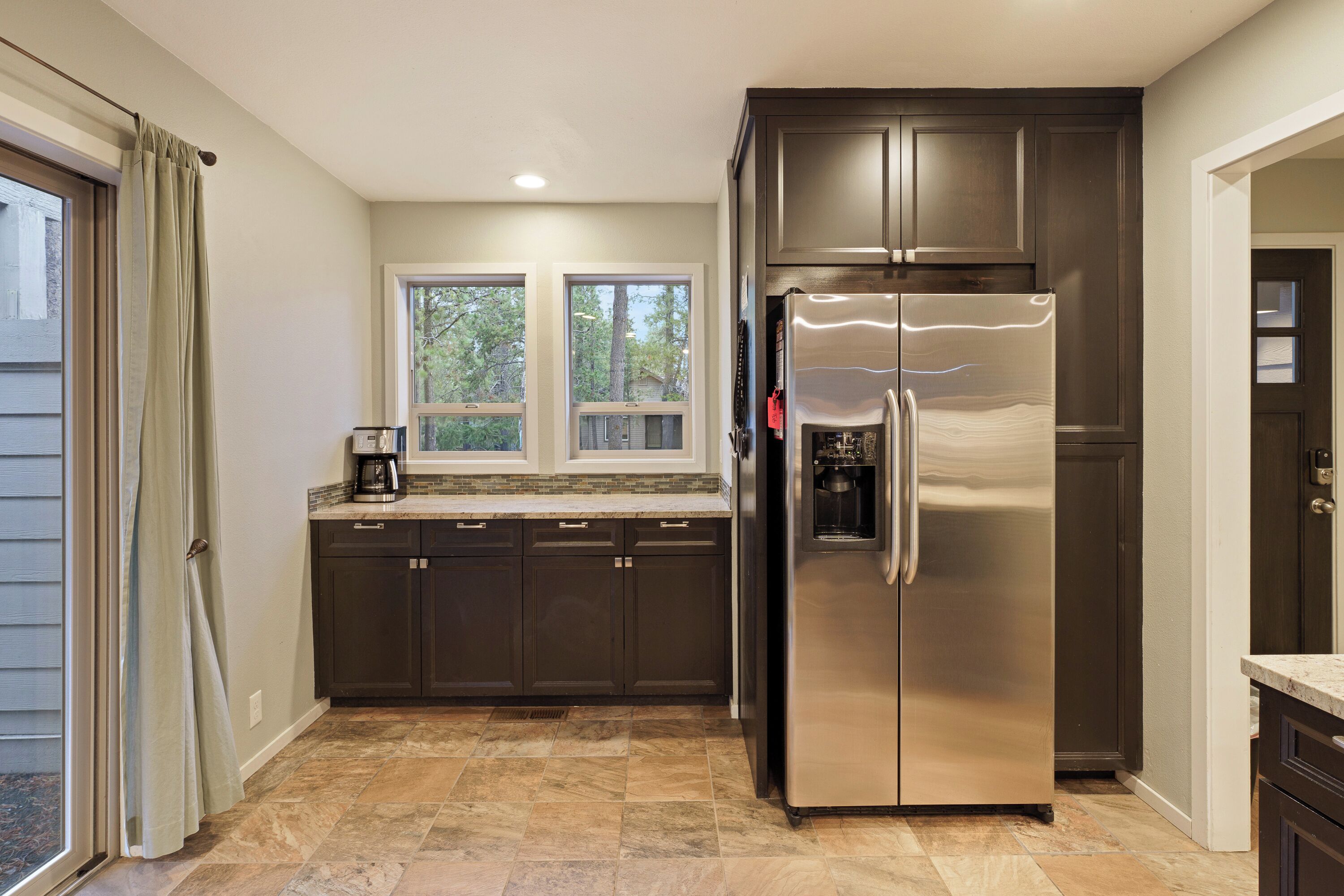 A kitchen with dark cabinets, a stainless steel refrigerator, a coffee maker, and a window with a view of trees.