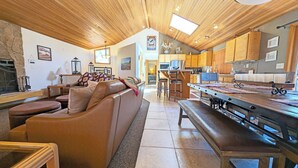 Een ruime woonkamer met houten plafond, een open haard, een bank en een eettafel met banken.