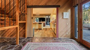 Un intérieur en bois avec un sol en pierre, une cuisine dotée d’un poêle en pierre et une vue sur l’extérieur à travers une porte vitrée.