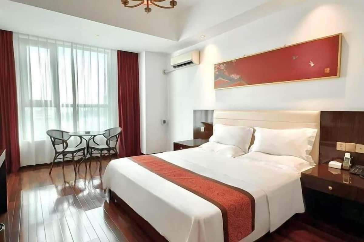 A hotel room with a large bed, a dining table with chairs, a desk with a telephone, and a large window with curtains.