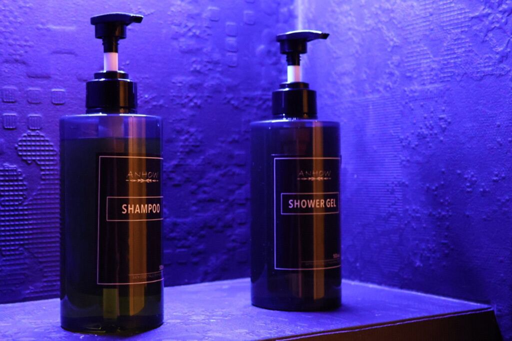 Deux bouteilles de shampoing et de gel douche Anhow sont posées sur une surface en bois, avec un mur texturé en arrière-plan.