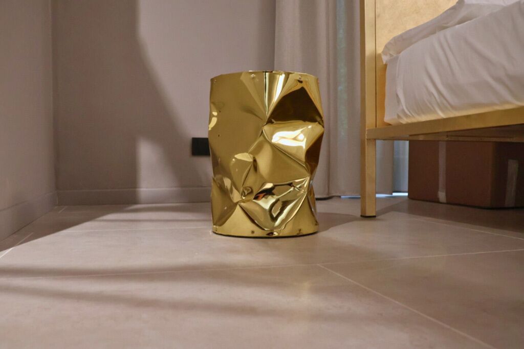 A modern room with a unique gold-colored cylindrical object on the floor, a bed with white bedding, and a plain wall.