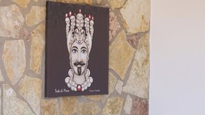 Une chambre avec un mur en pierre, un lit doté d’une tête de lit grise, une table de chevet blanche avec une lampe, et une œuvre d’art encadrée au-dessus du lit.