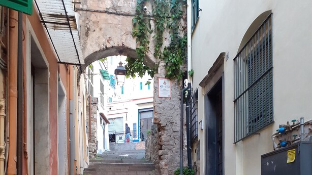 Une ruelle étroite, dallée de pierres et bordée de murs en pierre, qui mène à l’entrée d’un immeuble.