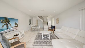 A modern living room with a white sofa, a coffee table, and a TV displaying a beach scene. A staircase leads to an upper level. - Intracostal Beauty in Fort Lauderdale (Fort Lauderdale)