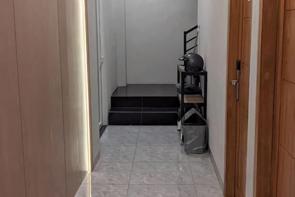 A narrow hallway with a wooden door on the right, a black shelf with items, and a trash bin. - SHAKTI SLEEP YOGYA (Ngampilan)