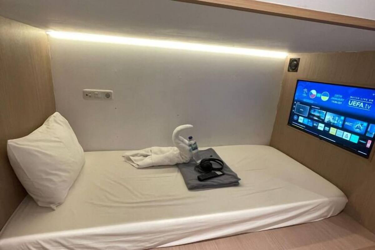 Ein Einzelbett mit einem weißen Kopfkissen, einem gefalteten Handtuch und einem Kopfhörerpaar. Ein flachbildfernseher an der Wand zeigt einen Sportkanal.
