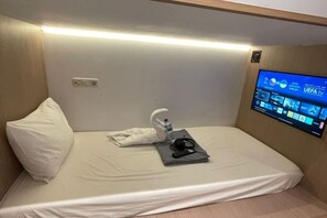 A single bed with a white pillow, a folded towel, and a pair of headphones on the bed. A flat-screen TV mounted on the wall displays a sports channel. - SHAKTI SLEEP YOGYA (Ngampilan)