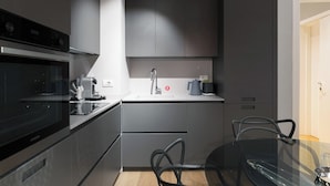 Une cuisine moderne avec des meubles gris, un four encastré, une machine à café et une table à manger en verre, accompagnée de chaises.