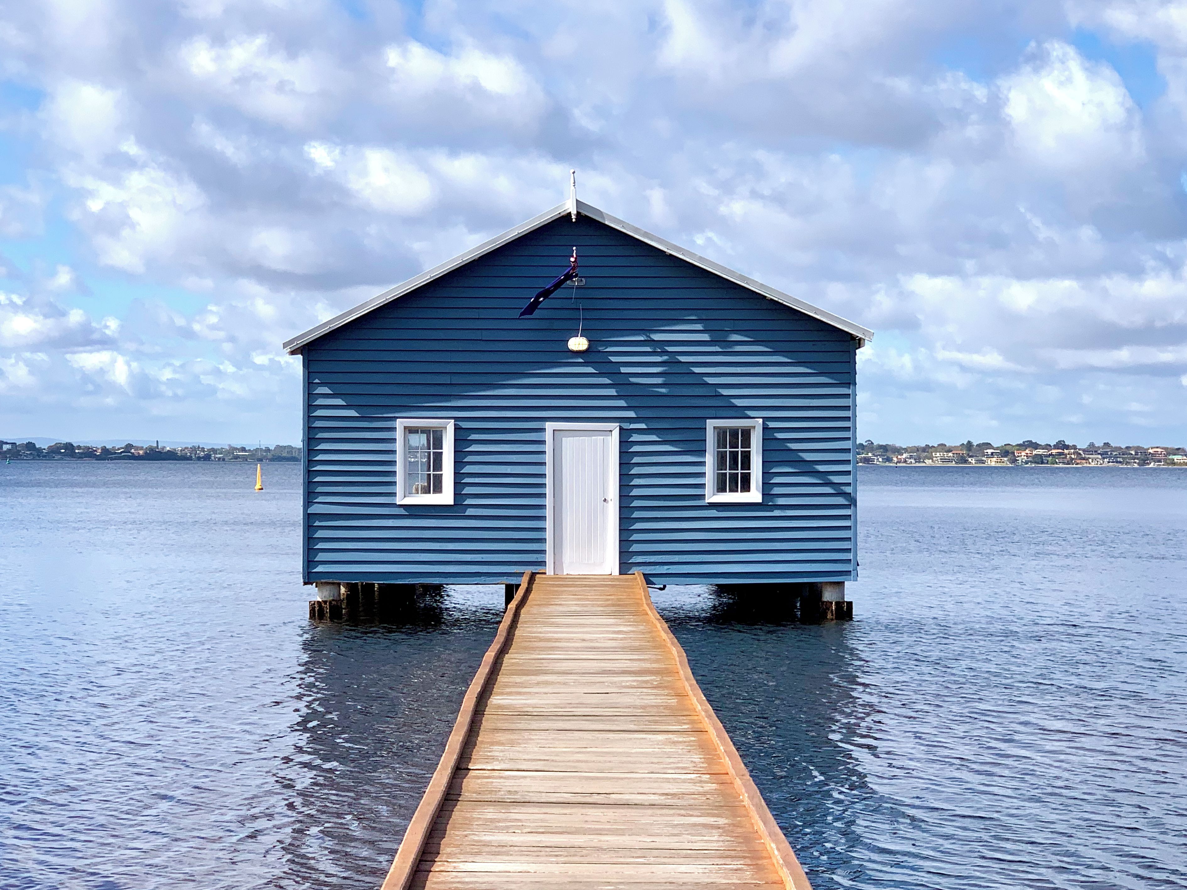 A blue wooden house with a white door and windows, connected to a wooden dock extending into calm water.