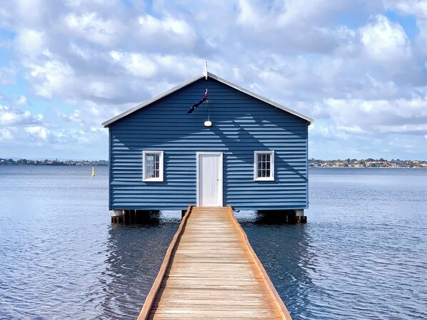 A blue wooden house with a white door and windows, connected to a wooden dock extending into calm water.