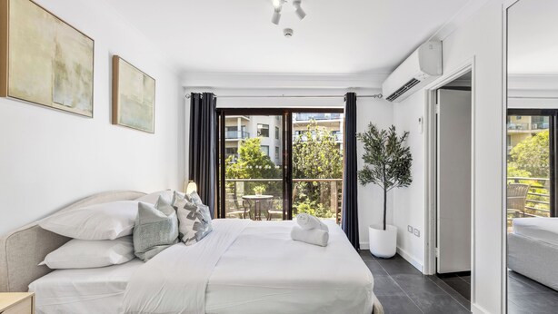 A modern bedroom with a large bed, a potted plant, and a balcony with a view of the city. - CBD Edge 1BR on Mounts Bay Road (Perth)
