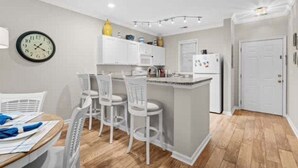 Una cocina moderna con armarios blancos, una isla central con taburetes de bar, un reloj en la pared y una puerta que da a otra habitación.