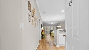 Un pasillo con piso de madera que conduce a una cocina con gabinetes blancos y un lámpara colgante.