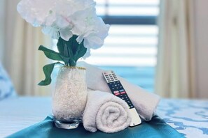 A bed with a blue and white patterned bedspread, a rolled white towel, a remote control, and a vase with white flowers. - The Bureau - Smart Suites Cyber (Cyberjaya)