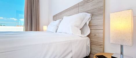 Habitación de hotel con una cama grande, una mesita de noche con lámpara y teléfono, y vista a la playa.
