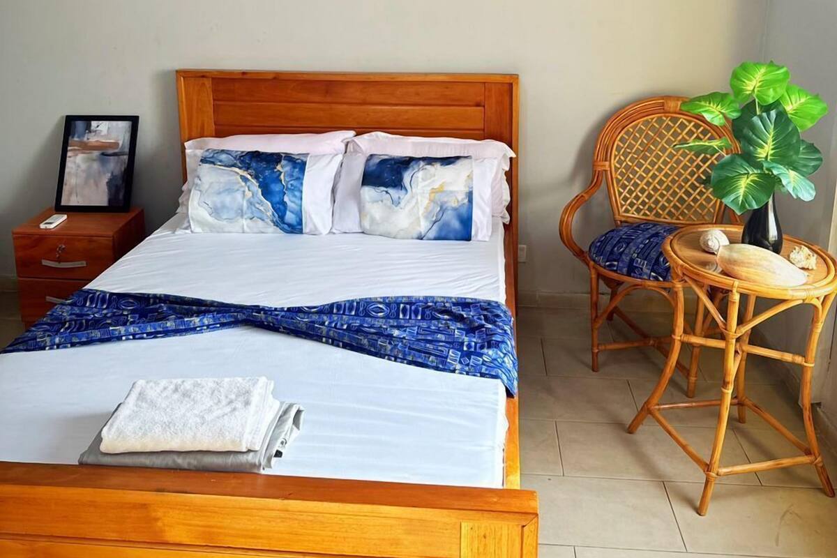 Une chambre avec un lit en bois, une table de chevet, une chaise en osier, une petite table avec une plante et un appareil de climatisation fixé au mur.