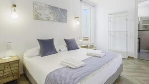 Une chambre d’hôtel moderne avec un grand lit, une table de chevet avec une lampe, un tableau au mur et une vue sur la cuisine par une porte ouverte.