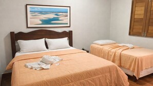 Ein Hotelzimmer mit einem großen Bett, einem kleineren Bett, einem Bild mit Strandmotiv und einem Fenster mit Holzjalousien.