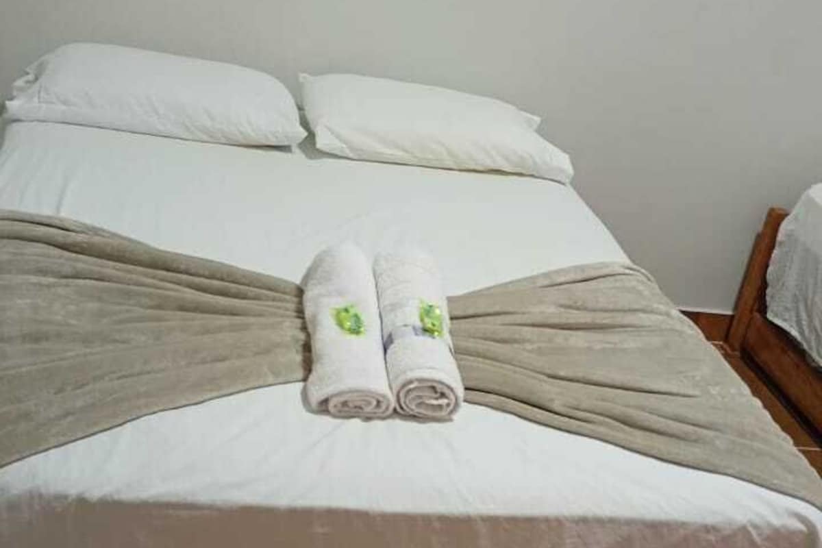 Una cama con una colcha blanca, dos toallas enrolladas con detalles verdes y una manta marrón.
