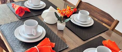 A dining table set with white cups and saucers, black placemats, and orange napkins.