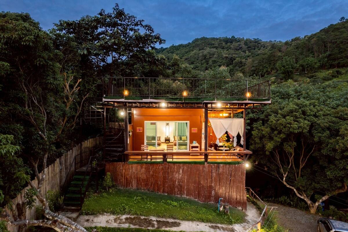 A two-story wooden cabin with a balcony, surrounded by dense forest.