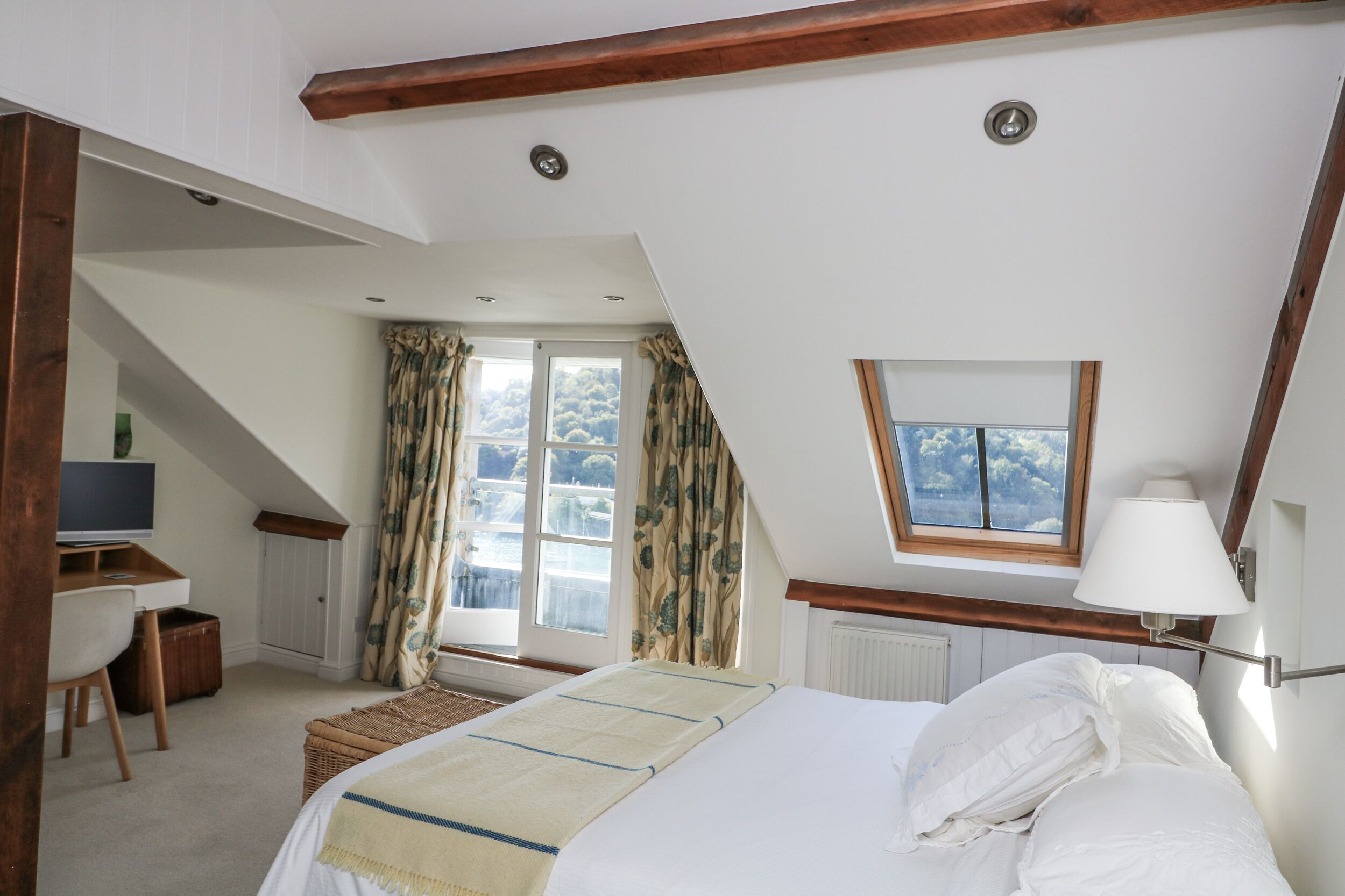 A bedroom with a sloped ceiling, a bed, a desk, a television, and a window with curtains.
