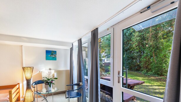 Une pièce moderne dotée d’une porte vitrée donnant sur un jardin, d’un banc en bois, d’une table en verre avec des chaises et d’un lampadaire.