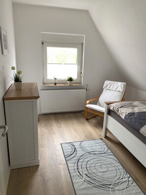 A small, well-lit room with a bed, a chair, a radiator, a window, and a small cabinet. - Huus an't Zingel - Holiday home in Krummhörn-Rysum (Krummhörn)