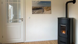 A room with a white door, a framed beach scene on the wall, and a black wood stove with a visible fire. - Huus an't Zingel - Holiday home in Krummhörn-Rysum (Krummhörn)
