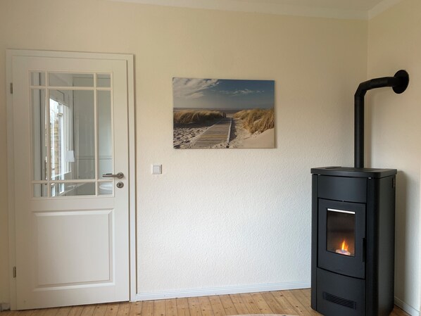 A room with a white door, a framed beach scene on the wall, and a black wood stove with a visible fire. - Huus an't Zingel - Holiday home in Krummhörn-Rysum (Krummhörn)
